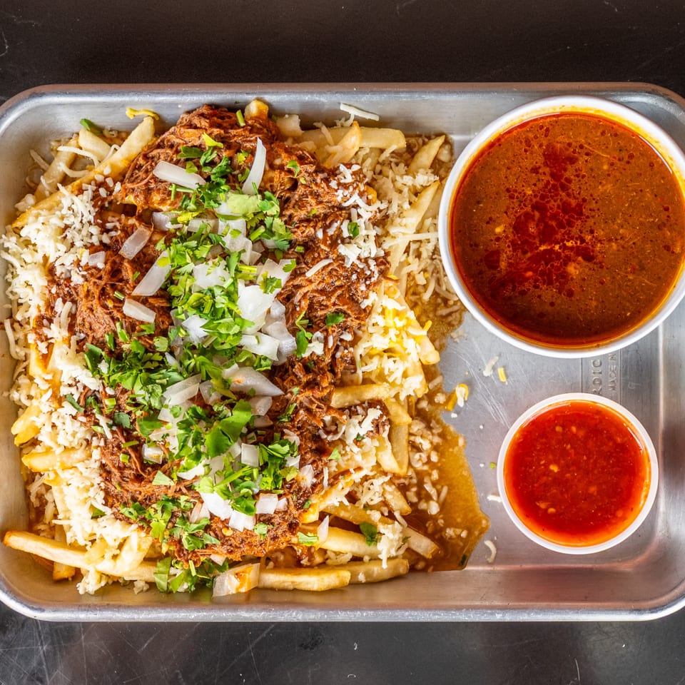 Birria fries with shredded beef, cheese, onions, cilantro, and two dipping sauces on a metal tray.