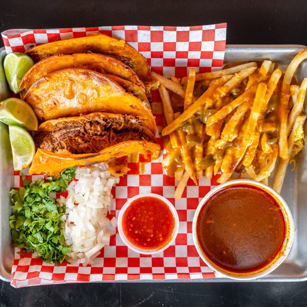 Birria tacos, chili cheese fries, cilantro, onions, red salsa, and consommé on a tray.