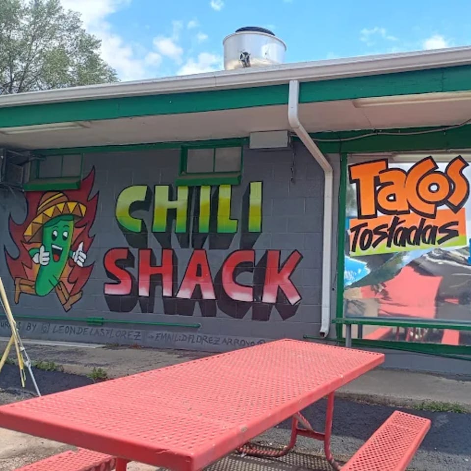 Exterior of Chili Shack with a red picnic table, mural of a chili pepper, and "Tacos Tostadas" sign.
