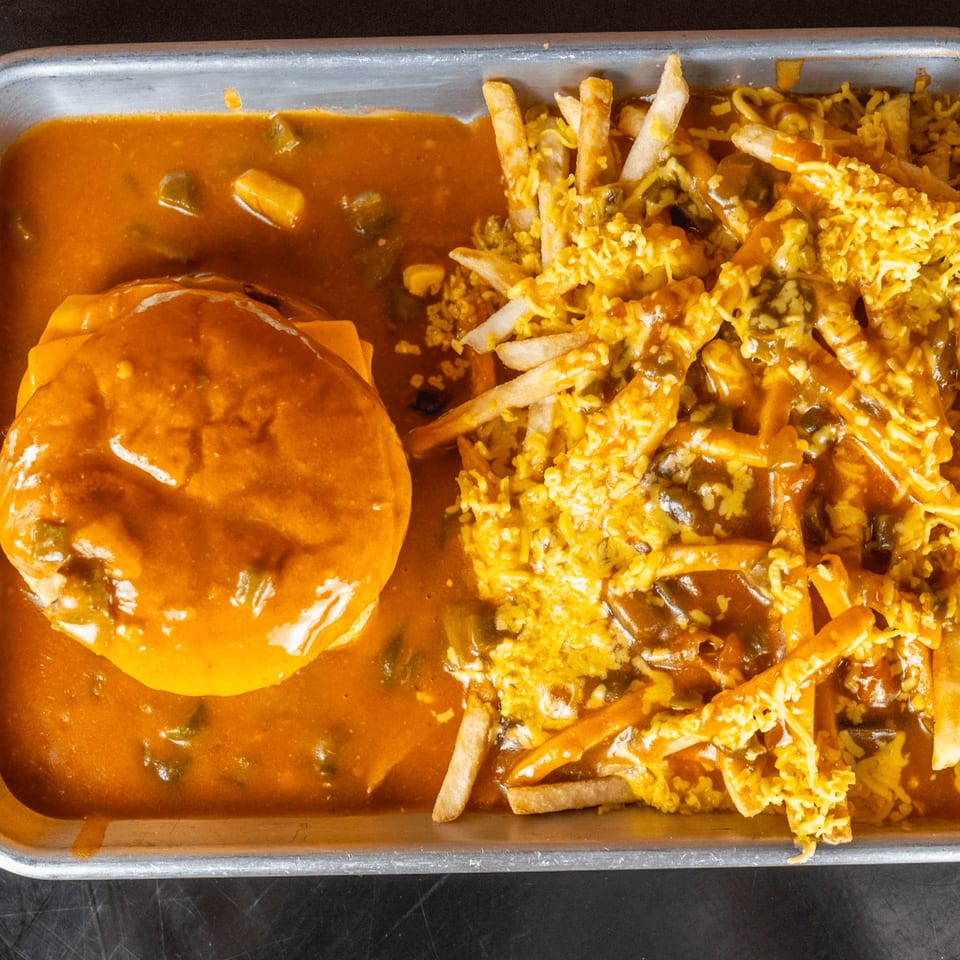 A burger covered in orange chili and cheese, next to chili cheese fries on a metal tray.