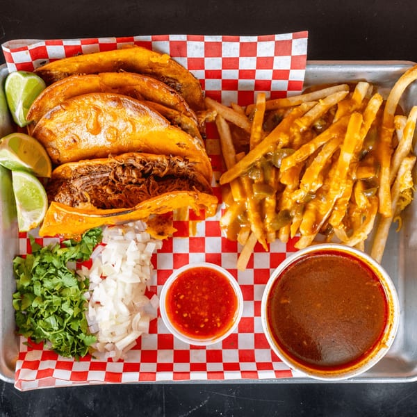 Birria tacos with limes, cilantro, onions, chili cheese fries, red salsa, and consommé on a tray.