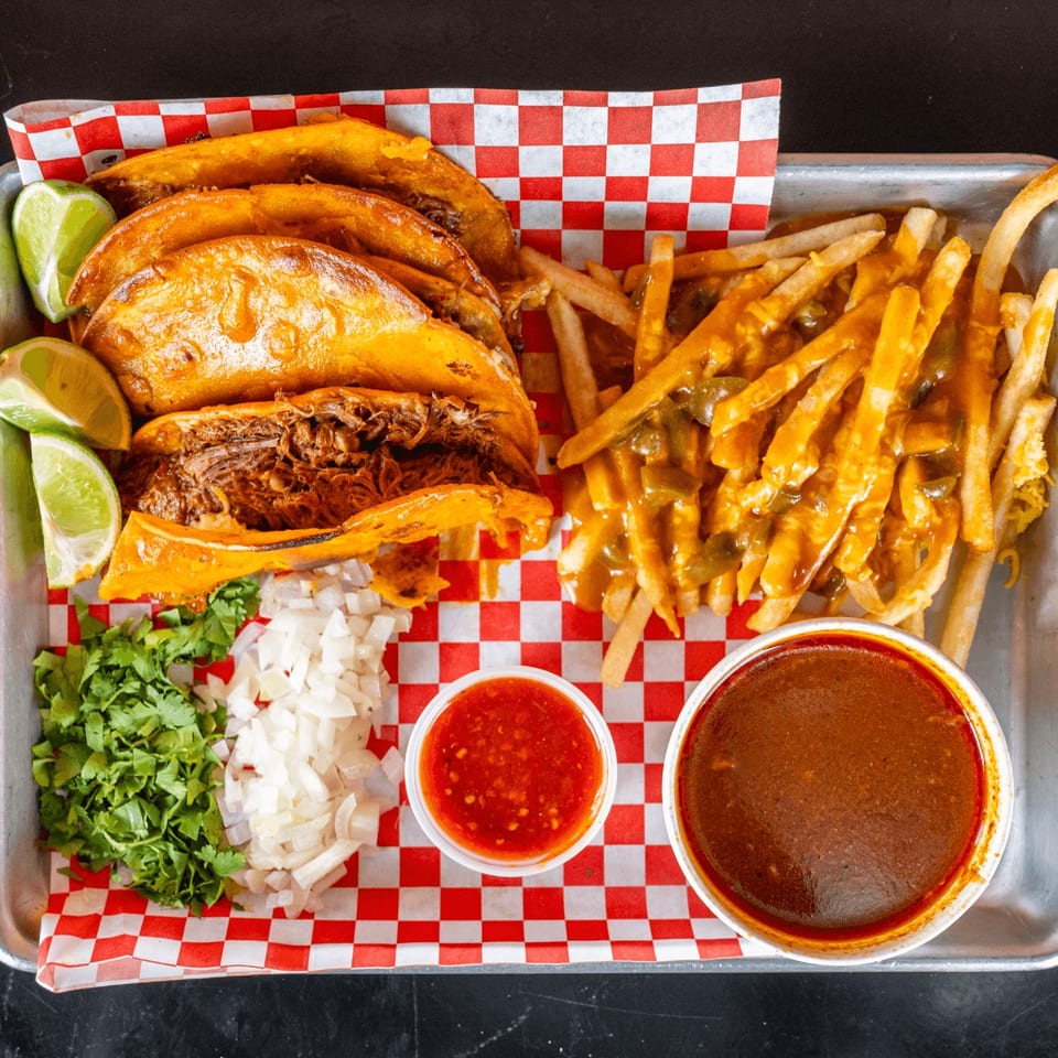 Birria tacos with limes, cilantro, onions, chili cheese fries, red salsa, and consommé on a tray.