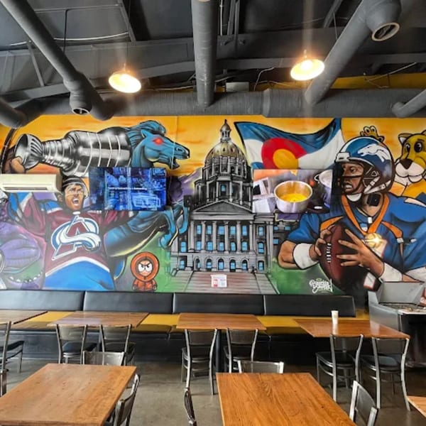 Restaurant interior with a vibrant mural featuring Colorado landmarks and sports figures above dining tables.
